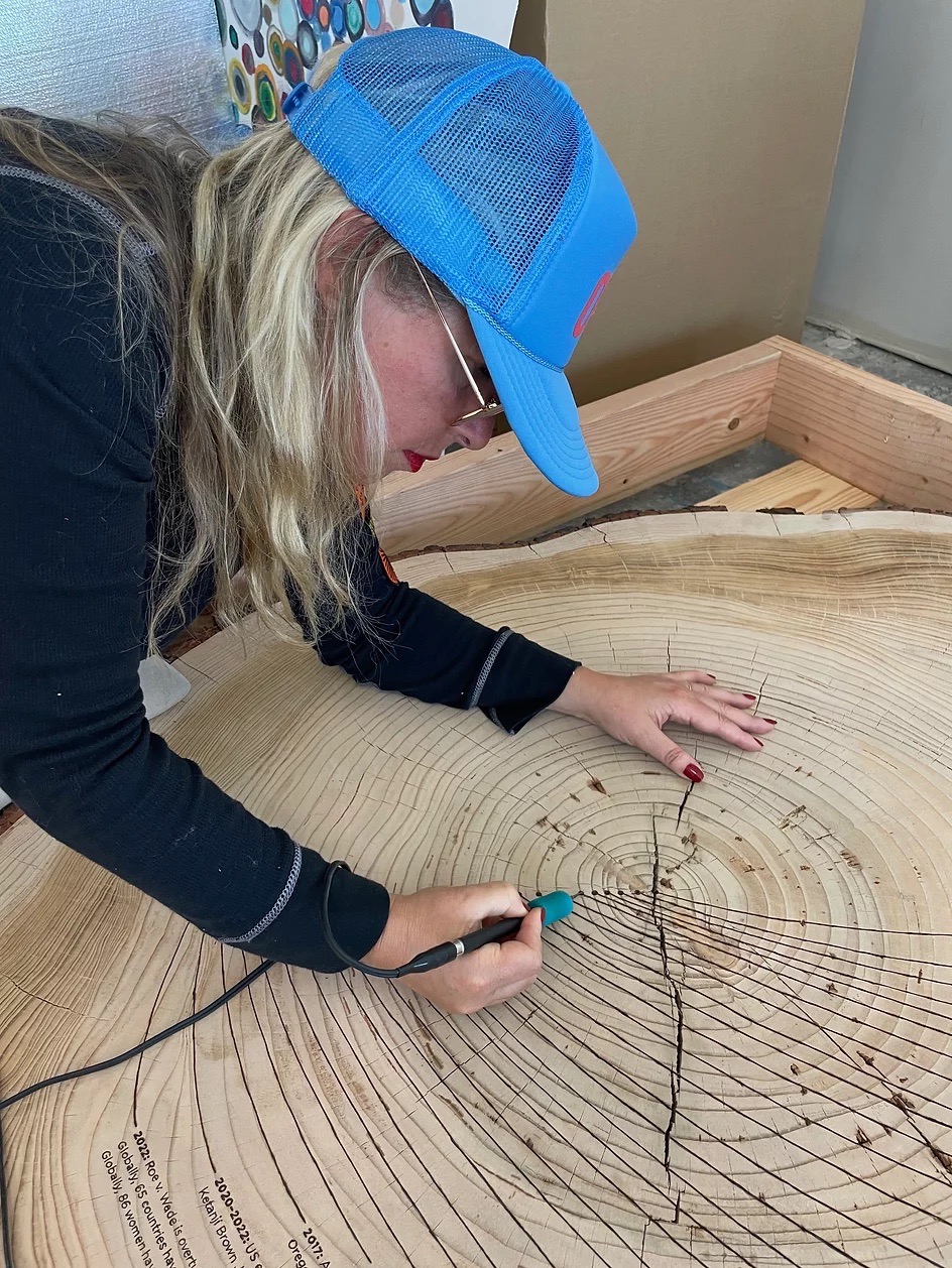 Tiffany Shlain in blue baseball cap leaning over to burn lettering into wood.