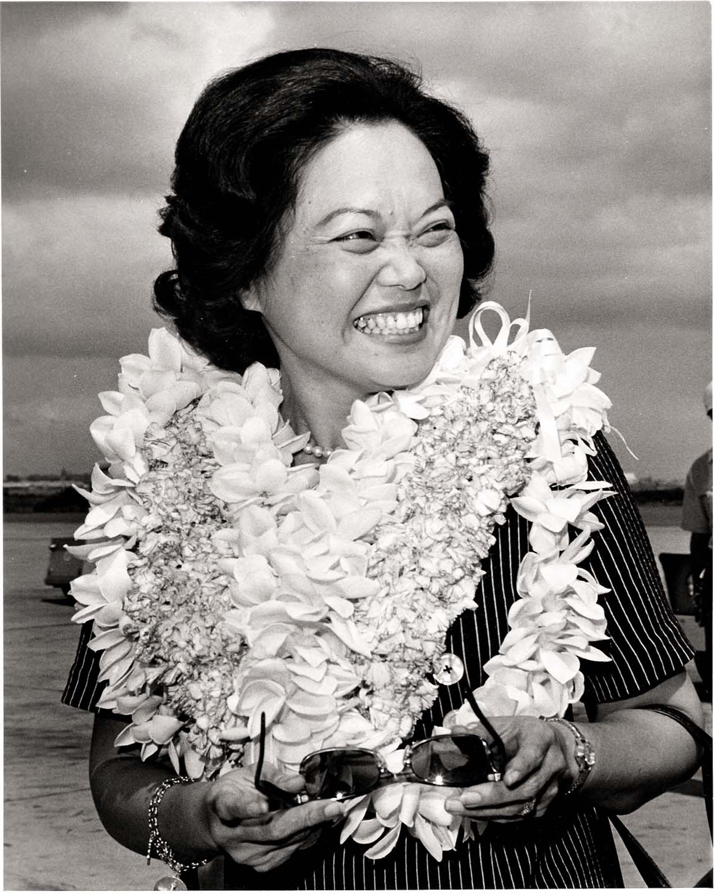 Black and white image of Patsy Mink wearing several Hawaiian leis and smiling.