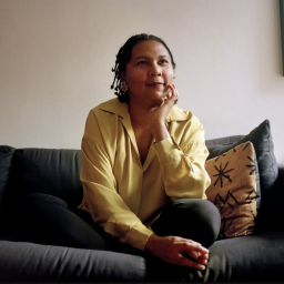 bell hooks sits on a green couch. She's wearing a yellow blouse, resting here chin on her hand, and staring off into the distance with look of hope. 