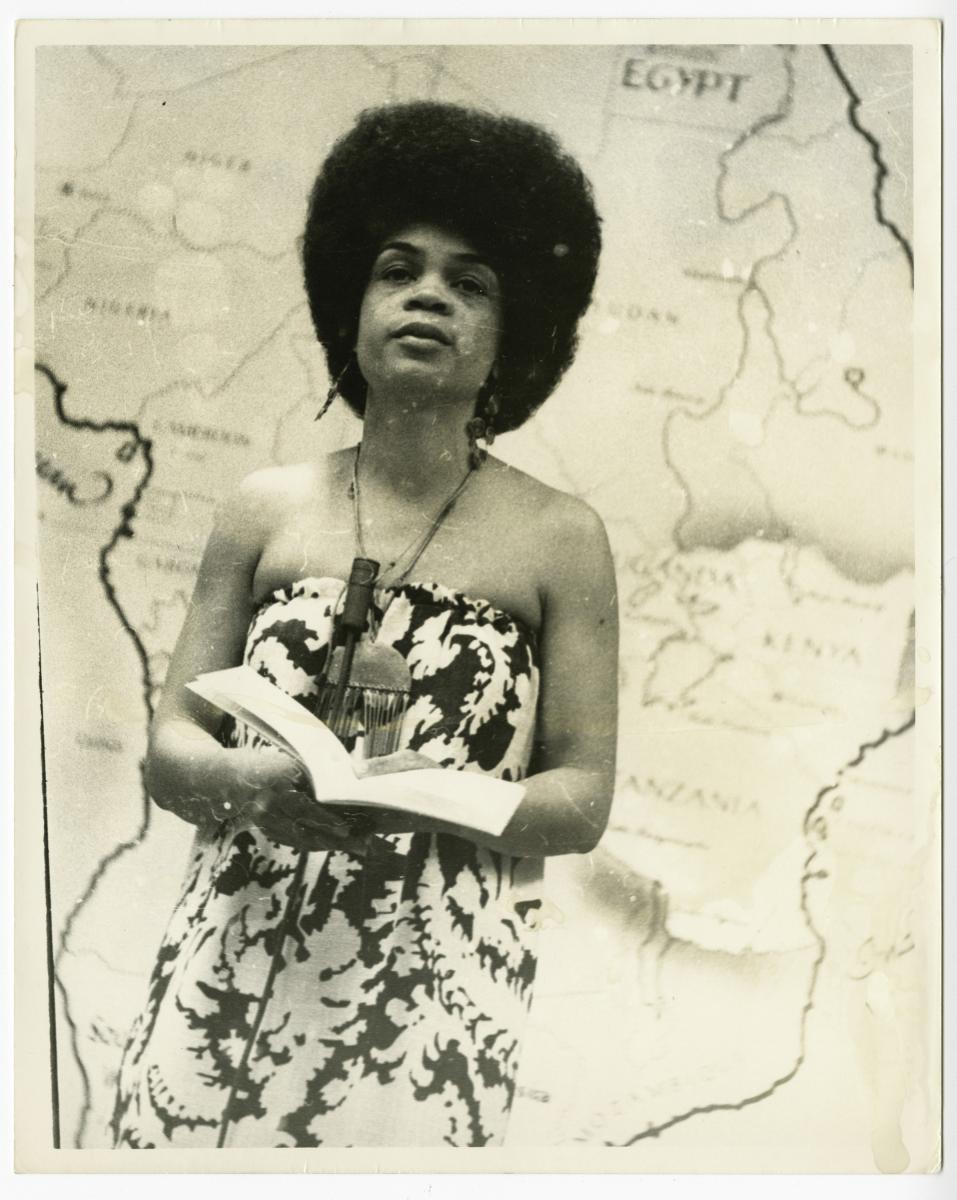 Photo of Sonia Sanchez stands holding a microphone and book poised to read poetry.