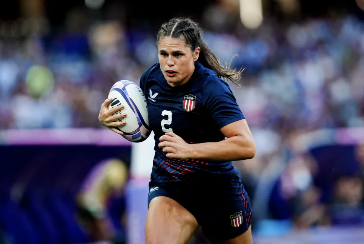 Woman in navy rugby uniform carries a rugby ball mid-play at the Paris Olympics