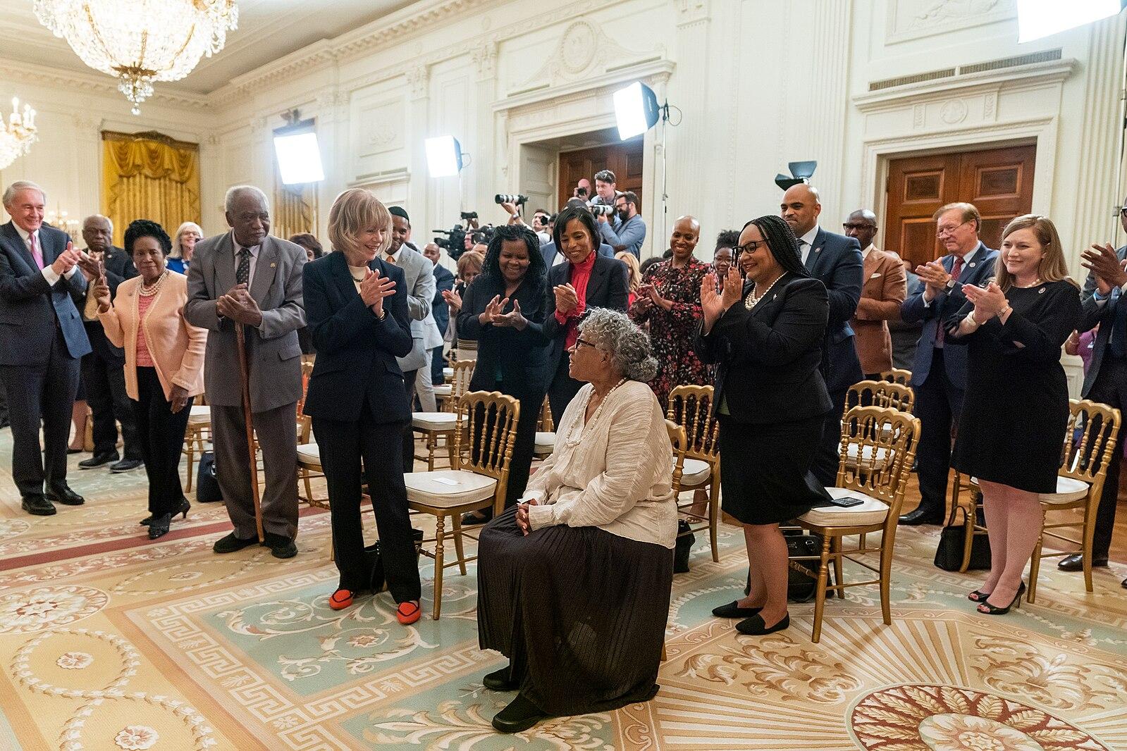 Opal Lee sits in a room while a crowd of people stand applauding her