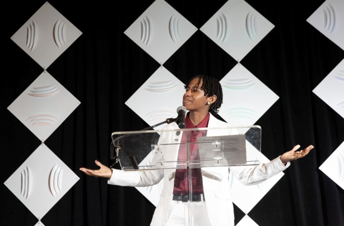 Kihana Wilson stands at a clear podium giving a talk.