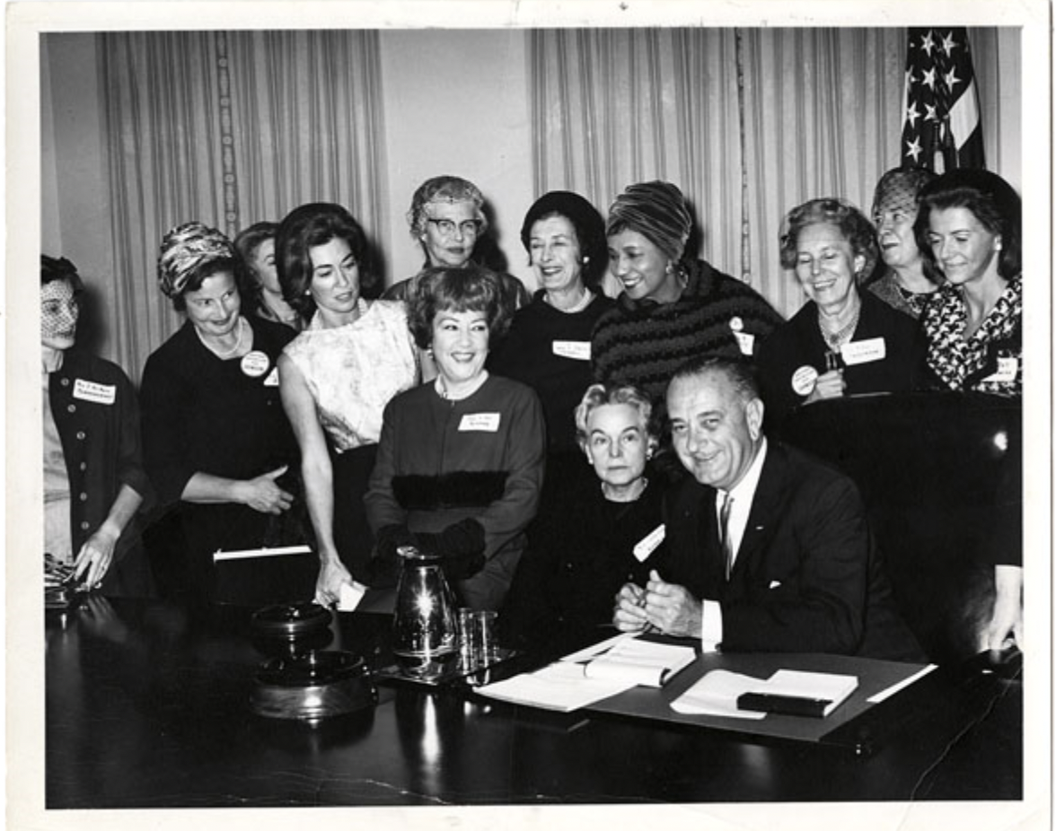 Dr. Helen Taussig sits beside President Lyndon B. Johnson at a desk surrounded by others.