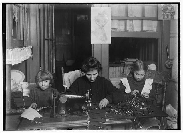 Black & white photo of a family of two children along with their mother seated at a table with an old sewing machine.