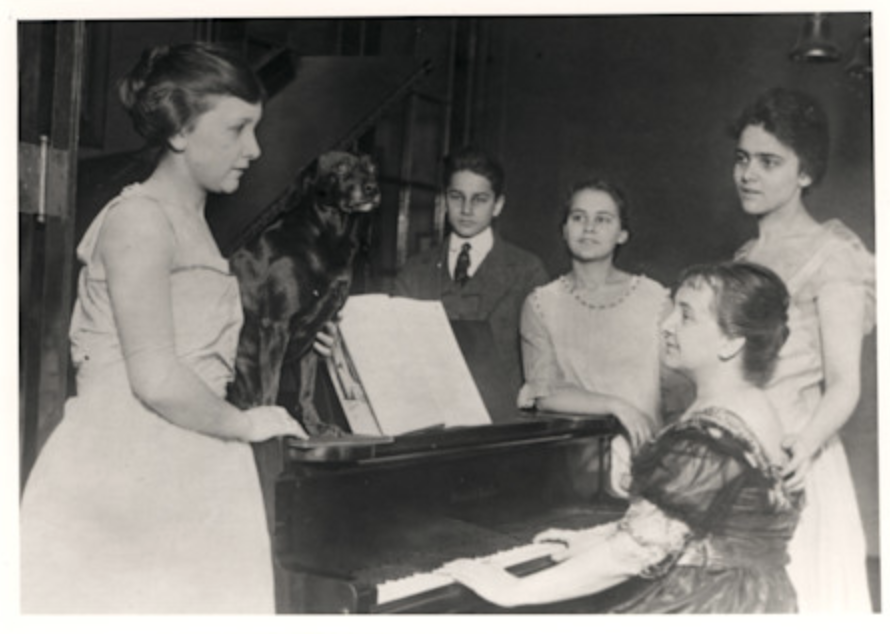 A family stands around a photo with one person seated playing the piano