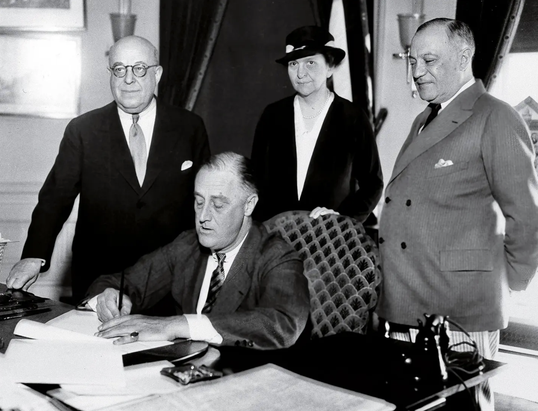 Black and white photo of Perkins standing behind President Roosevelt as he signs a document at his desk.