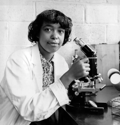 Black and white photo of Bath in a lab coat posing with a microscope.