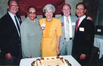 Color photo of five people, Cooke-Wright included that are posing with a cake celebrating Cooke-Wright