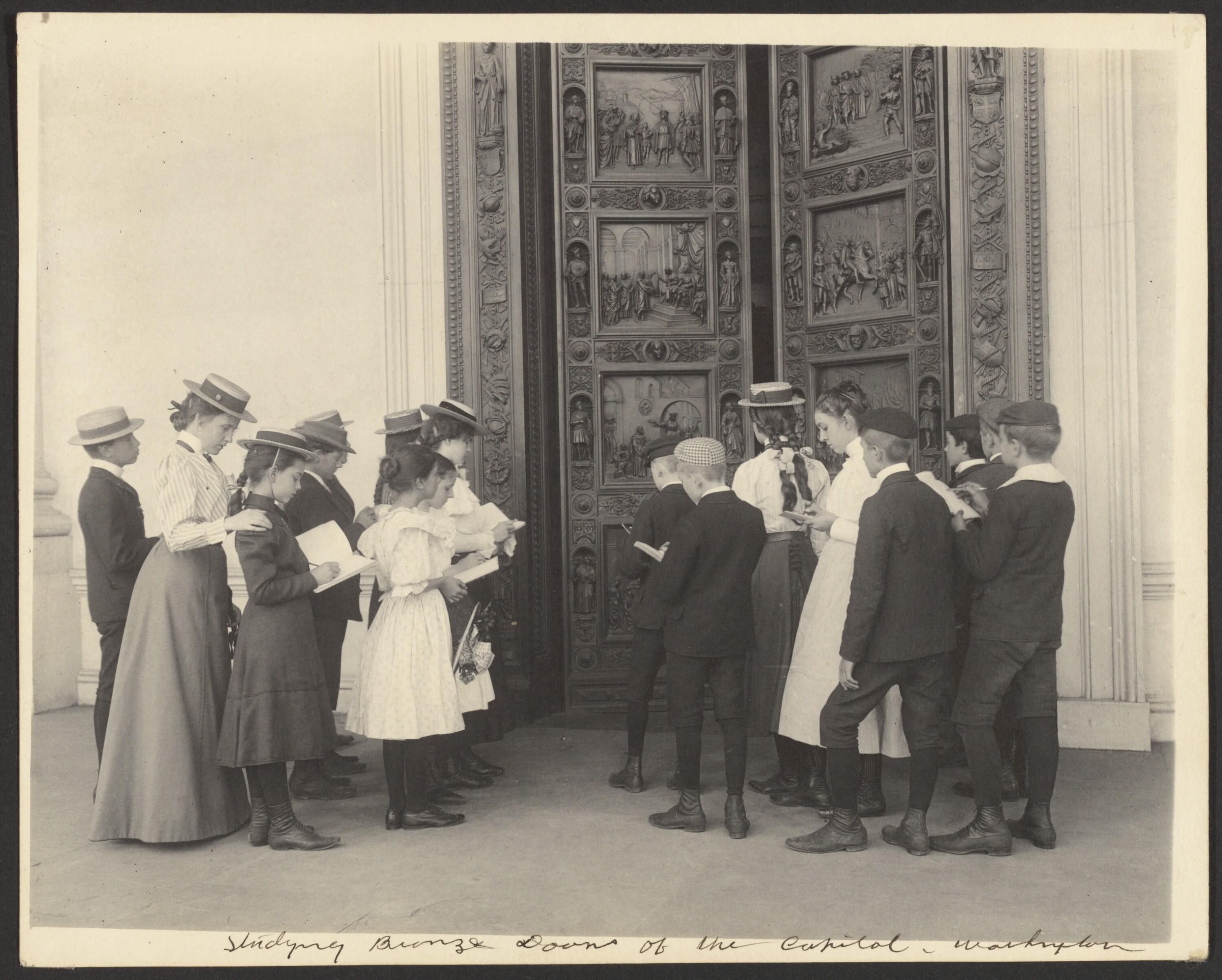 Black and white photo of elementary age students on a field trip. They are standing in front of a large door that features many carvings and are taking notes/sketching on notebooks. 