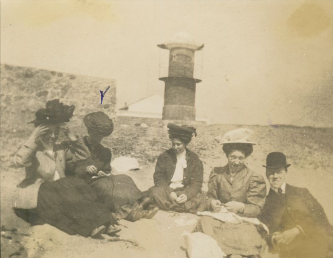 Aged photograph of five people sitting on the beach with a lighthouse-like structure behind them. They all wear decorative hats and formal attire. 
