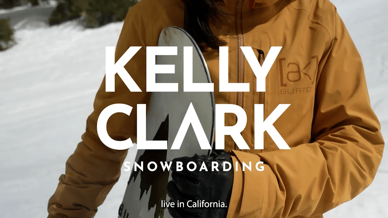 Promotional picture with text "Kelly Clark Snowboarding" in front of Clark, who is wearing dark yellow gear and holding a snowboard.