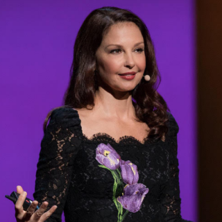 Image of Ashley Judd speaking. She wears a black dress with purple flowers and stands in front of a purple background.