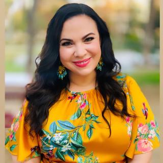 Gloria Calderón Kellett, smiling, seated in yellow dress with colorful flowers and yellow and turquoise earrings.