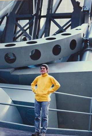 Photo of young Andrea Ghez, wearing a yellow sweater, standing in front large space equipment.