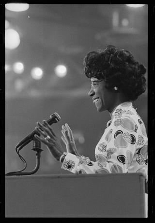 Black and white photo of Shirley Chisholm behind a podium, smiling and waving to the audience. The photo is of her left profile.