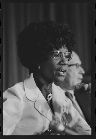 Black and white photo of Shirley Chisholm speaking into a microphone, facing to the left.