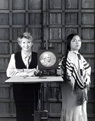 Black and white photo of Goodacre standing with Randy'L Teton, with the coin design in between them. Goodacre is smiling, and Teton is posing like the coin.