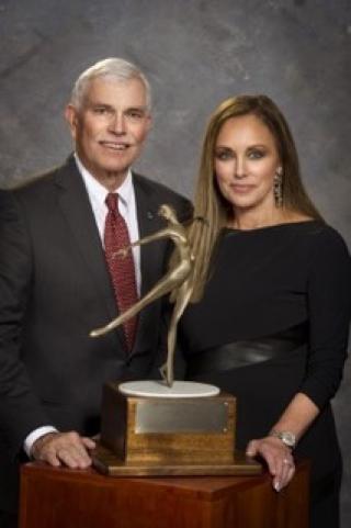 Photo of Peggy Fleming standing behind a large trophy. She is accompanied by a man in a suit.