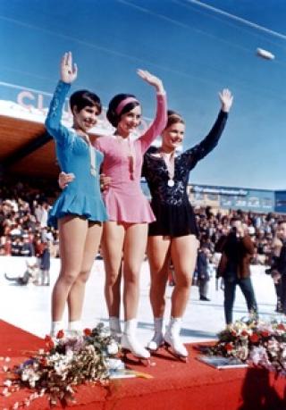 Photo of Peggy Fleming (center) with her first World Gold Medal. There is one woman on each side of Fleming. All three are waving and smiling. 