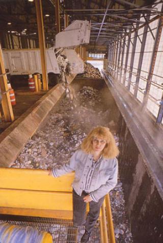 Photo of Ukeles standing in a garbage transfer station in front of collected garbage. She is looking up at the camera.