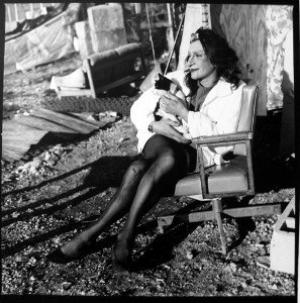 A black and white photograph of Sylvia Rivera wearing black tights and white jacket, sitting in a chair with a cat in her lap.
