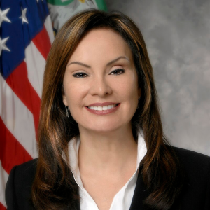 Headshot of Rosie Rios in black blazer with white shirt in front of American flag posted to her right.