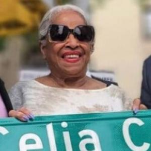 Ruth Sánchez, wearing sunglasses, holds a sign that reads "Celia Cruz"