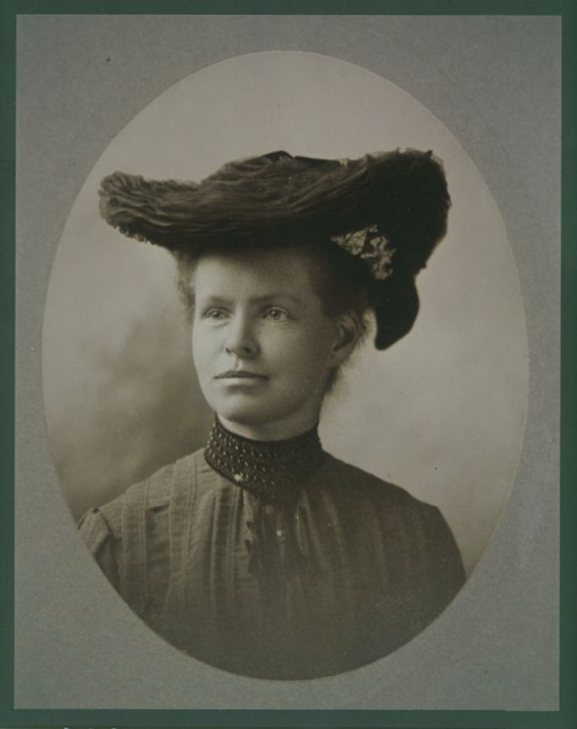 Portrait of Nettie Stevens. She wears a large and decorative hat while staring into the distance. 