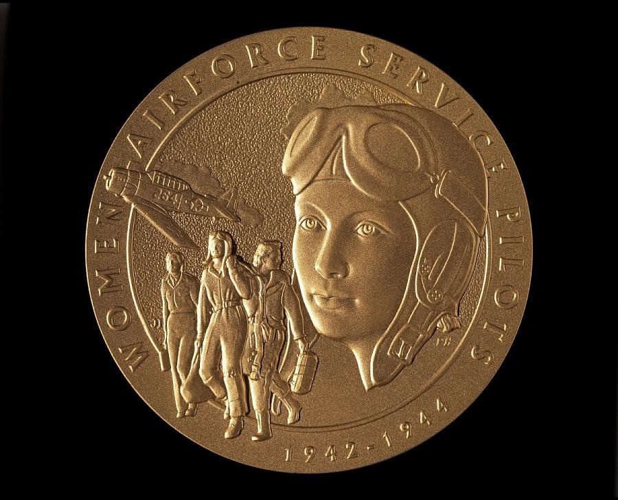 Photograph of the WASP Medal, a gold medal with an engraving of a woman pilot's face, three other women pilots, and a plane.