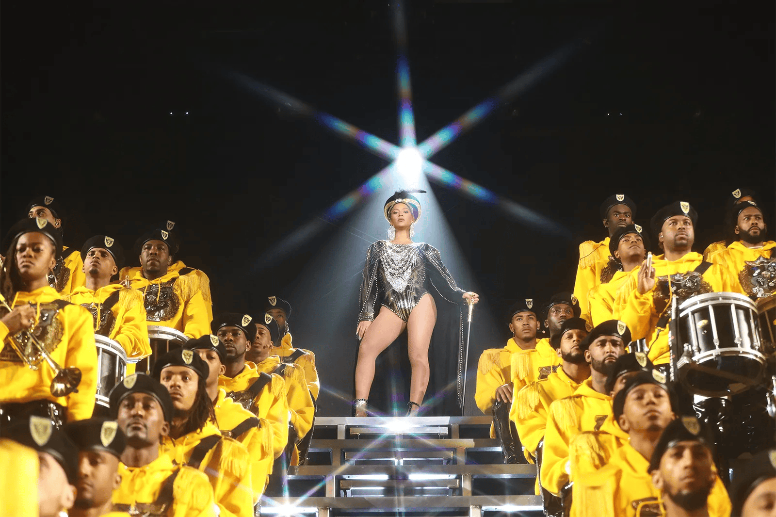 Photo of Beyonce (center) performing on a raised stage. A spotlight shines behind her, and there are groups of people on each side of Beyonce.