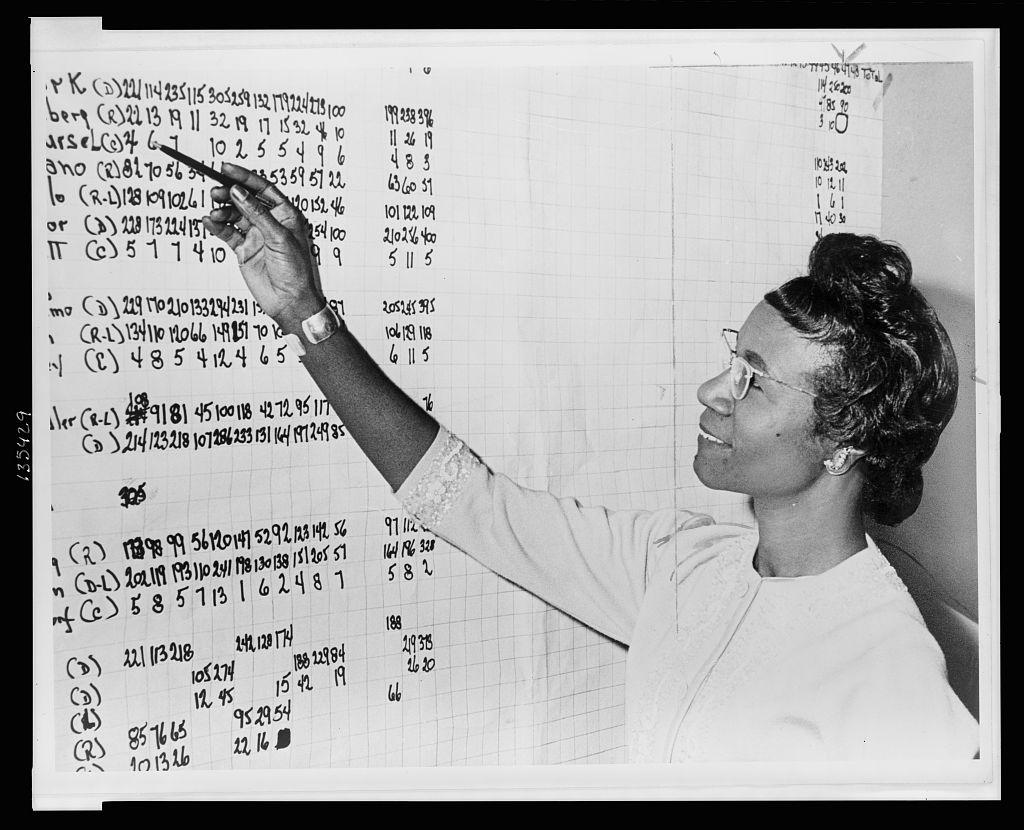 Black and white photo of young Chisholm looking at a board of poll numbers, pointing her pen at the top of the board.