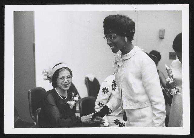Black and white photo of Shirley Chisholm shaking Rosa Parks hand. Chisholm is standing in a white suit and Parks is sitting.