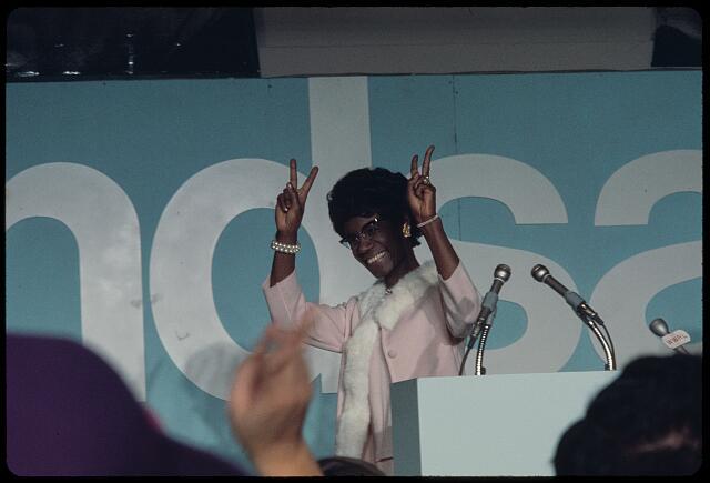 Photo of Shirley Chisholm on the campaign trail. She is behind a podium, smiling and holding up "Peace" signs.