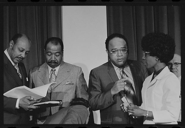 Black and white photo of Shirley Chisholm standing amongst members of the Congressional Black Caucus. Chisholm is on the far right of the image, with three men to the left.