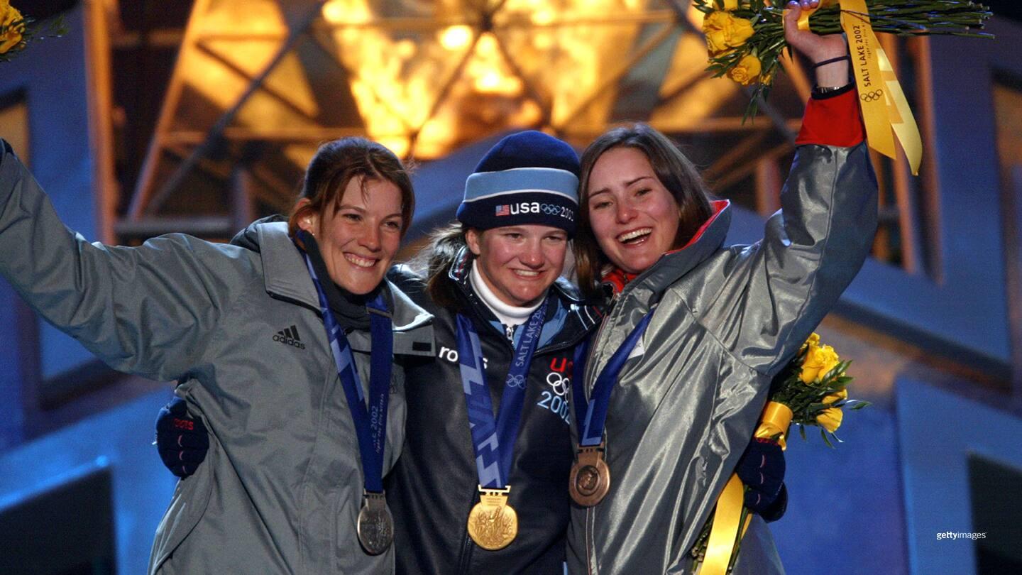 Kelly Clark, wearing a gold medal, stands in the middle of two other women with silver and bronze medals. All three are smiling and celebrating.