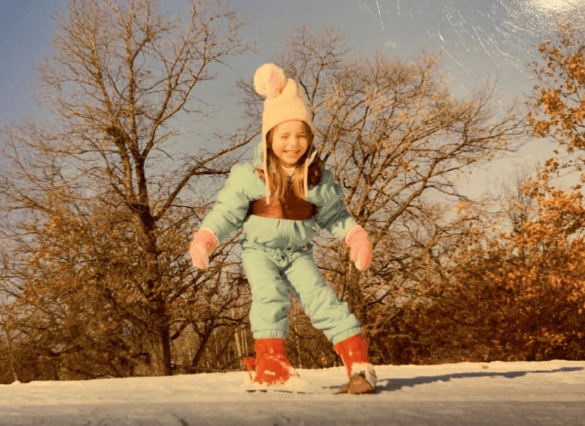 Photo of young Lindsey Vonn in winter gear, wearing a large white beanie. She is smiling.