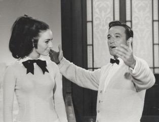 Photo of Peggy Fleming looking at a man with his arms open. She is wearing a white shirt with a large black bow.