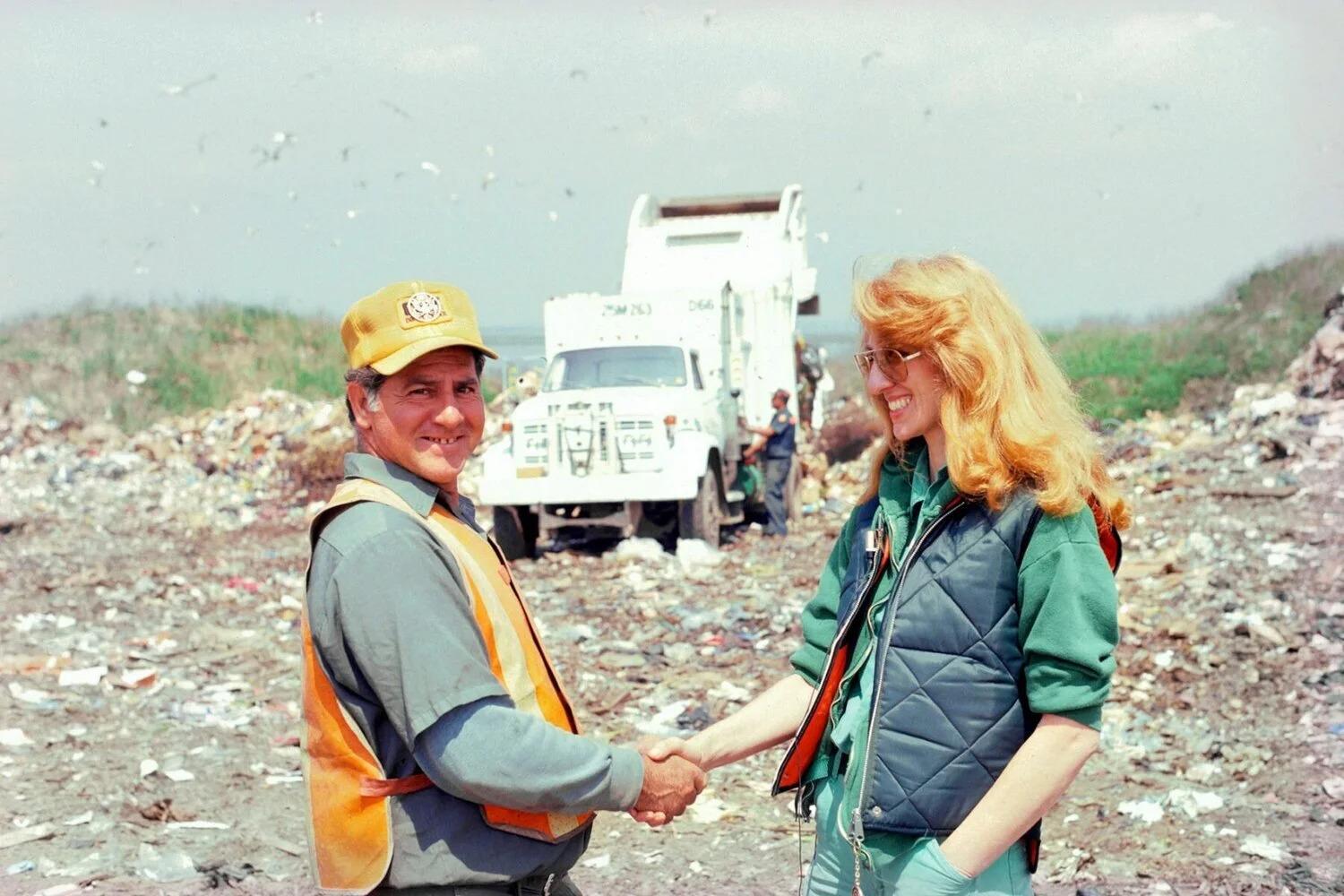 Photo of Ukeles (right) shaking hands with a sanitation worker in a landfill. Ukeles is looking at the worker, who is looking at the camera. Both are smiling.
